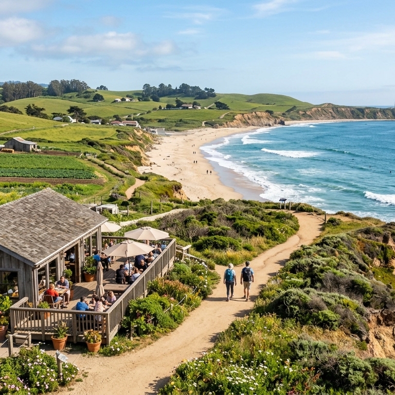 An aerial view of a rustic wooden restaurant with a outdoor patio sitting on a coastal cliff, next to a walking trail, farm fields, sandy beach, and lighthouse in the distance.